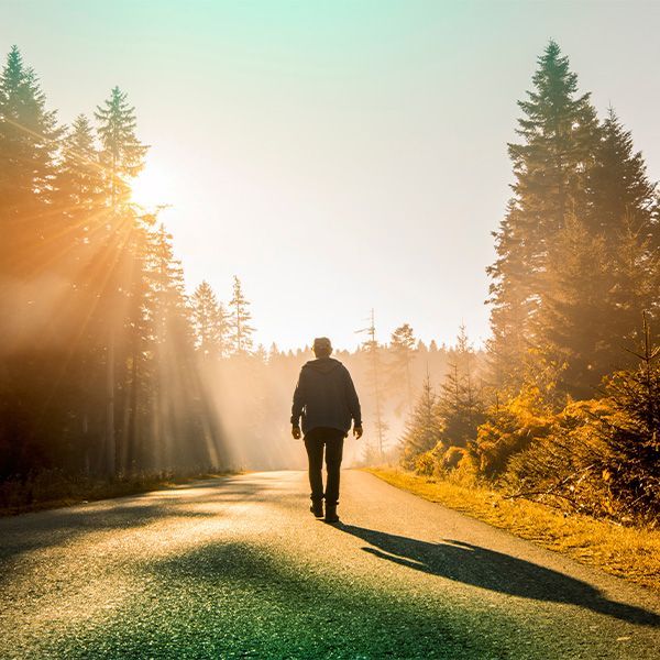 A man is walking down a road in the woods at sunset.